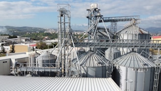 A complex industrial facility with multiple large metal silos and various interconnected pipes and structures, set against a backdrop of distant hills and a clear sky. The surrounding area includes other industrial buildings and green landscapes.