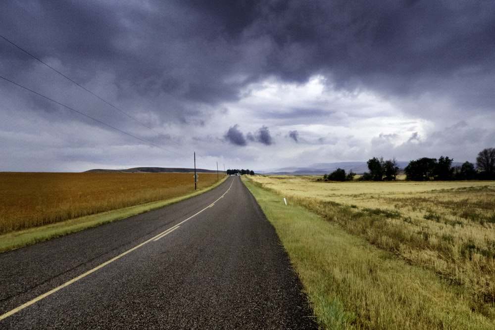 Route goudronnée grise entre un champ d’herbe brune sous un ciel nuageux ensoleillé bleu et blanc pendant la journée