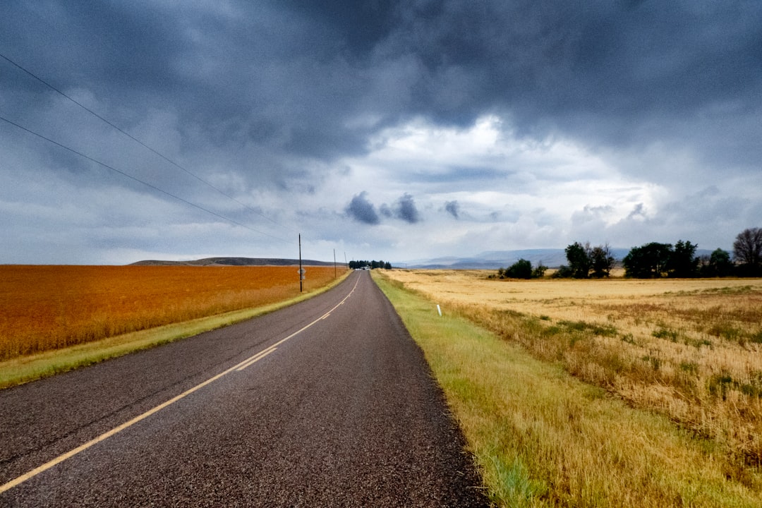 gray asphalt road between brown grass field under blue and white sunny cloudy sky during daytime