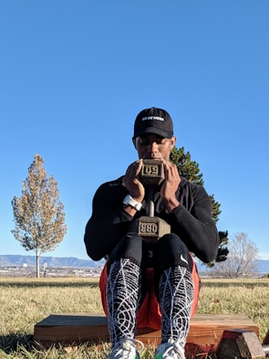 A person is sitting outdoors on a wooden block, holding a weight close to their chest. They are wearing a black hat, long-sleeve shirt, patterned leggings, and sneakers. The background shows a clear blue sky, a couple of trees, and distant mountains.