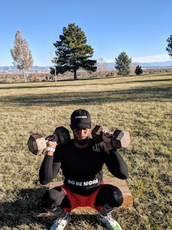 A person is exercising outdoors, holding two dumbbells while squatting. The individual is wearing a black long-sleeved shirt, red shorts, and a black cap with the words 'GO BE MORE' on both the shirt and cap. The background features a grassy field with several trees, and mountains can be seen in the distance.