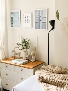 A cozy and stylish room corner features a wooden dresser with a light-colored top. On the dresser are decorative items including a vase with flowers, a wooden horse figurine, two stuffed toys, and several neatly stacked books. The wall above the dresser is adorned with three framed artworks, and a black floor lamp stands to the right. A knitted beige blanket is draped over a bed, contributing to the warm and inviting atmosphere.