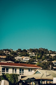 A scenic view featuring a hillside neighborhood with several houses nestled among dense green trees. The sky above is a clear, rich blue, creating a vivid contrast. Roofs of the houses vary, with some in red and others in gray, reflecting a mix of architectural styles.