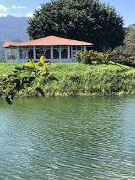 A serene landscape featuring a small white house with a red-tiled roof, surrounded by lush greenery. The house is located on a grassy hill adjacent to a calm body of water with gentle ripples. Large trees provide a natural backdrop, and mountains are visible in the distance under a clear blue sky.