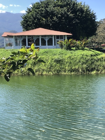 A serene landscape featuring a small white house with a red-tiled roof, surrounded by lush greenery. The house is located on a grassy hill adjacent to a calm body of water with gentle ripples. Large trees provide a natural backdrop, and mountains are visible in the distance under a clear blue sky.