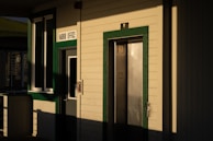 A building exterior with a sign reading 'Harbor Office' above a door. The building features green trim around windows and doors, and there is an elevator door next to the office door. The angle of the sunlight casts long shadows across the facade.