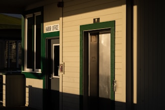 A building exterior with a sign reading 'Harbor Office' above a door. The building features green trim around windows and doors, and there is an elevator door next to the office door. The angle of the sunlight casts long shadows across the facade.