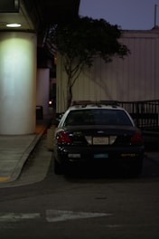 A police car is parked next to a large white pillar. The car is positioned on the side of a building with a tree beside it, and the area appears to be dimly lit with streetlights. There is no visible activity around the car, and the overall environment seems quiet.