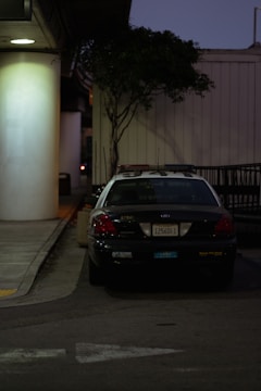 A police car is parked next to a large white pillar. The car is positioned on the side of a building with a tree beside it, and the area appears to be dimly lit with streetlights. There is no visible activity around the car, and the overall environment seems quiet.