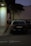 A police car is parked next to a large white pillar. The car is positioned on the side of a building with a tree beside it, and the area appears to be dimly lit with streetlights. There is no visible activity around the car, and the overall environment seems quiet.