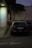 A police car is parked next to a large white pillar. The car is positioned on the side of a building with a tree beside it, and the area appears to be dimly lit with streetlights. There is no visible activity around the car, and the overall environment seems quiet.