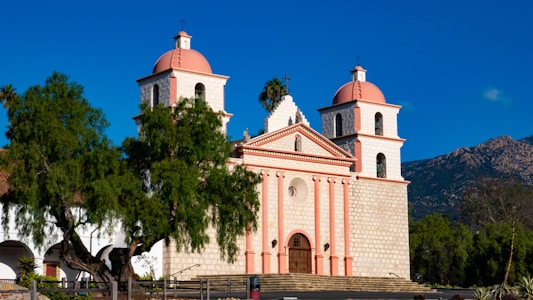 A historic mission-style church with two bell towers and a central facade, featuring a pink and beige color scheme. The building is surrounded by lush green trees with a mountain backdrop under a clear blue sky.