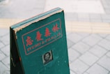 A green, weathered signboard with red lettering and Japanese characters, featuring the words 'STUDIO & FILM LAB.' There is a small, vintage black-and-white portrait near the bottom of the sign. The sign is standing on a tiled pavement.