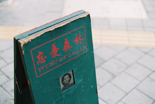 A green, weathered signboard with red lettering and Japanese characters, featuring the words 'STUDIO & FILM LAB.' There is a small, vintage black-and-white portrait near the bottom of the sign. The sign is standing on a tiled pavement.