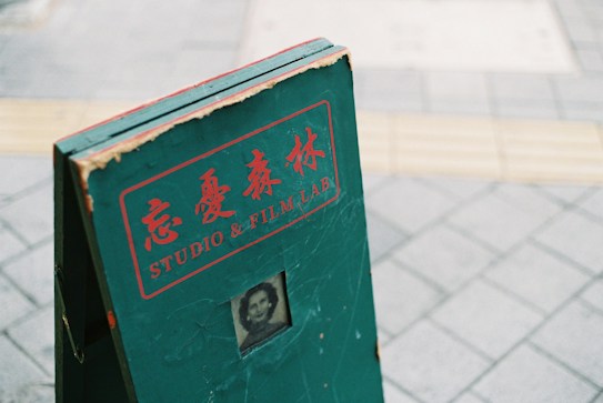 A green, weathered signboard with red lettering and Japanese characters, featuring the words 'STUDIO & FILM LAB.' There is a small, vintage black-and-white portrait near the bottom of the sign. The sign is standing on a tiled pavement.