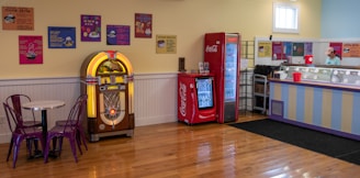 A retro-themed indoor setting featuring a jukebox, Coca-Cola vending machines, a round table with two purple chairs, and an ice cream counter. The walls are decorated with colorful vintage posters. The floors are wooden and clean.