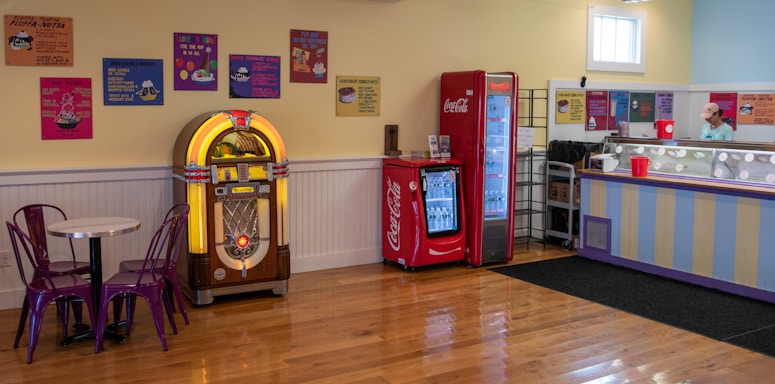 A retro-themed indoor setting featuring a jukebox, Coca-Cola vending machines, a round table with two purple chairs, and an ice cream counter. The walls are decorated with colorful vintage posters. The floors are wooden and clean.