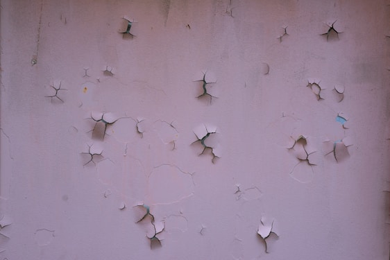 A textured wall with multiple areas of cracked and peeling paint, revealing darker surfaces underneath.
