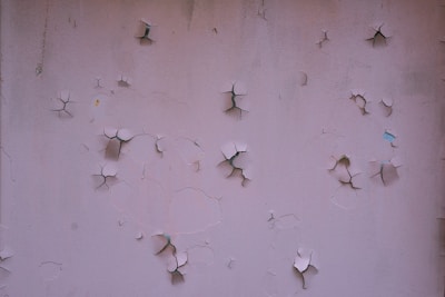 A textured wall with multiple areas of cracked and peeling paint, revealing darker surfaces underneath.