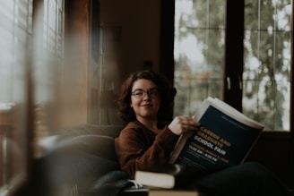 A person with glasses sits comfortably with a book open in their hands, suggesting a relaxed reading moment. The setting appears cozy, with soft lighting coming through large windows, creating a calm atmosphere.