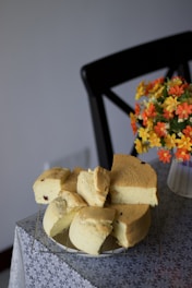 A sponge cake, cut into several pieces, is resting on a wire rack placed on a table covered with a patterned tablecloth. Beside the cake, there is a vase containing vibrant artificial flowers in shades of orange and yellow. A black chair is partially visible in the background.