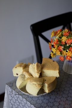 A sponge cake, cut into several pieces, is resting on a wire rack placed on a table covered with a patterned tablecloth. Beside the cake, there is a vase containing vibrant artificial flowers in shades of orange and yellow. A black chair is partially visible in the background.