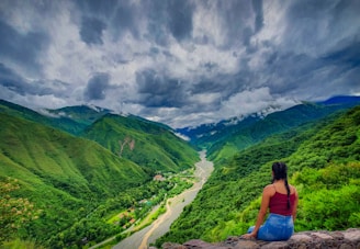 woman in blue tank top sitting on rock near green mountains under white clouds during daytime
