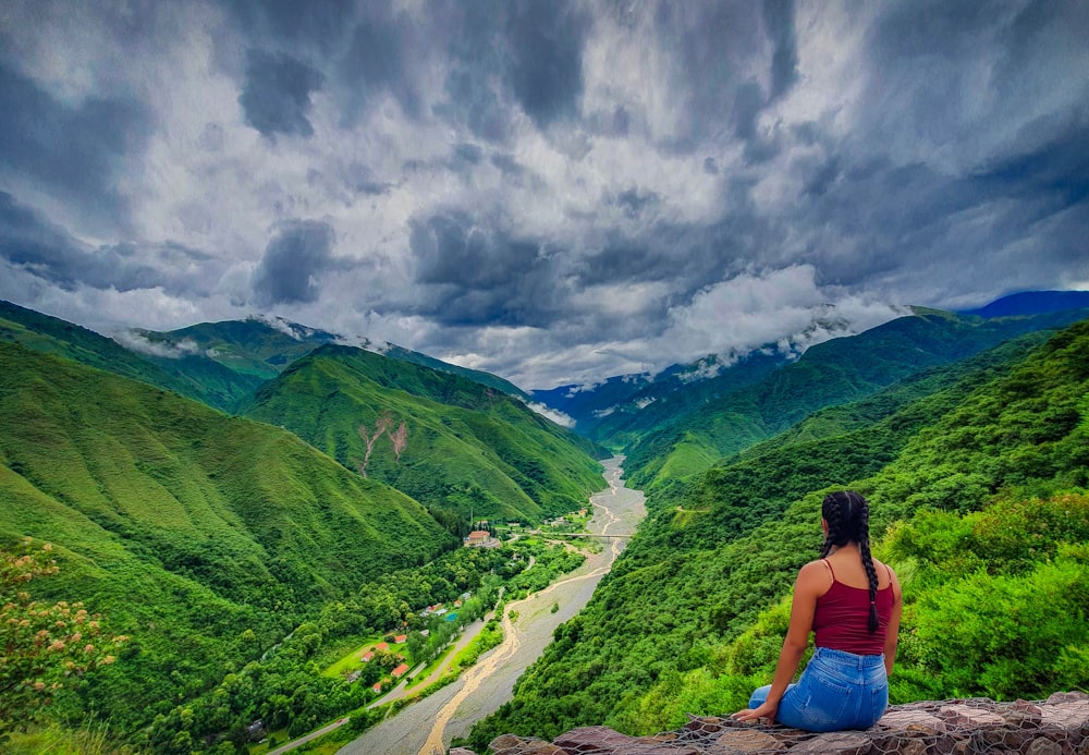 woman in blue tank top sitting on rock near green mountains under white clouds during daytime