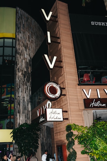 A restaurant facade displaying prominent signage with the word 'VIVO' and 'italian kitchen'. The exterior features a mix of wood and concrete textures, with lights illuminating the area. Greenery surrounds the entrance, and several people are gathered near the doorway.
