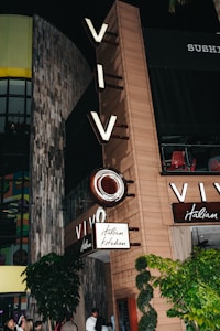 A restaurant facade displaying prominent signage with the word 'VIVO' and 'italian kitchen'. The exterior features a mix of wood and concrete textures, with lights illuminating the area. Greenery surrounds the entrance, and several people are gathered near the doorway.