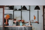 A cozy cafe kitchen features two people engaged in tasks, one wearing a yellow beanie, the other a cap. Shelves hold black teapots, cups, and a toaster oven, while a mint green weighing scale and stacks of bowls occupy the counter. Industrial black pendant lights hang overhead, creating a warm, inviting ambiance.