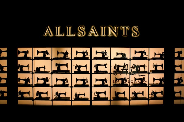 A storefront display features a series of vintage sewing machines, each placed in its own illuminated square compartment. Above the display, the word 'ALLSAINTS' is prominently displayed in gold lettering. The arrangement creates a striking visual pattern with a focus on the silhouette of each sewing machine.