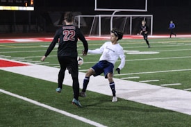 Two soccer players, one in a black uniform labeled 'Titans', and the other in a white top and blue shorts, are engaging in a match on a well-lit artificial turf field at night. The player in white is positioned defensively, attempting to block or intercept the ball from the opponent. The background includes a soccer goal and a few other players and figures, with a brightly illuminated scoreboard displaying scores.