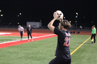 A soccer player wearing a black jersey with the number 21 is preparing to throw the ball from the sideline. The field is a combination of green artificial turf and red track material, illuminated by floodlights in a nighttime setting. Several other players and a referee are visible in the background.
