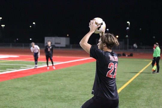 A soccer player wearing a black jersey with the number 21 is preparing to throw the ball from the sideline. The field is a combination of green artificial turf and red track material, illuminated by floodlights in a nighttime setting. Several other players and a referee are visible in the background.