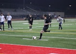 Under the lights of a stadium at night, a soccer player in a black uniform is about to kick a ball on an artificial turf field. Several other players in white and black uniforms are standing and watching. There are bleachers in the background with few spectators.