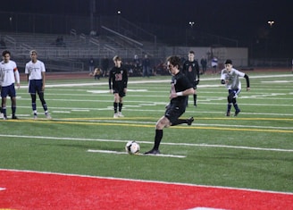 Under the lights of a stadium at night, a soccer player in a black uniform is about to kick a ball on an artificial turf field. Several other players in white and black uniforms are standing and watching. There are bleachers in the background with few spectators.