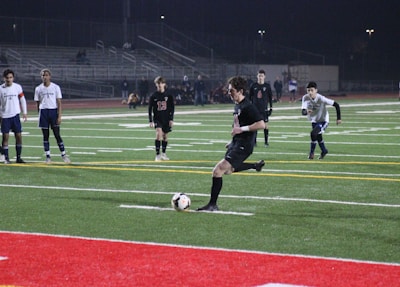 Under the lights of a stadium at night, a soccer player in a black uniform is about to kick a ball on an artificial turf field. Several other players in white and black uniforms are standing and watching. There are bleachers in the background with few spectators.