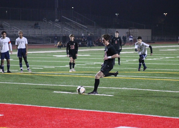 Under the lights of a stadium at night, a soccer player in a black uniform is about to kick a ball on an artificial turf field. Several other players in white and black uniforms are standing and watching. There are bleachers in the background with few spectators.