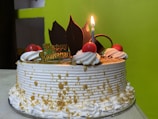 A decorated cake with white frosting is adorned with red cherries and chocolate pieces. A lit candle stands on top, next to a small sign that reads 'Happy Anniversary'. The cake has decorative icing swirls and gold sprinkles.
