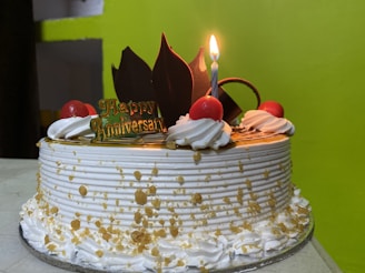 A decorated cake with white frosting is adorned with red cherries and chocolate pieces. A lit candle stands on top, next to a small sign that reads 'Happy Anniversary'. The cake has decorative icing swirls and gold sprinkles.