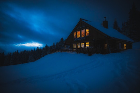 A cabin surrounded by snow-covered ground, with illuminated windows emitting a warm glow, contrasting against the dark, cloudy night sky. Pine trees line the horizon, adding depth to the wintry scene.