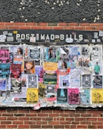 An outdoor bulletin board attached to a brick wall is covered with various colorful posters and flyers. These posters feature information about music events, concerts, and other local activities. The board appears weathered, with some posters overlapping and corners peeling from exposure.