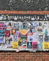 An outdoor bulletin board attached to a brick wall is covered with various colorful posters and flyers. These posters feature information about music events, concerts, and other local activities. The board appears weathered, with some posters overlapping and corners peeling from exposure.