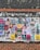 An outdoor bulletin board attached to a brick wall is covered with various colorful posters and flyers. These posters feature information about music events, concerts, and other local activities. The board appears weathered, with some posters overlapping and corners peeling from exposure.
