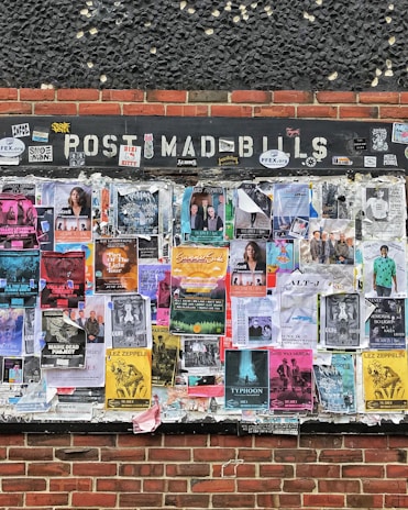 An outdoor bulletin board attached to a brick wall is covered with various colorful posters and flyers. These posters feature information about music events, concerts, and other local activities. The board appears weathered, with some posters overlapping and corners peeling from exposure.