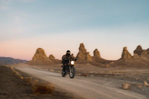 A dusty dirt road stretching into a golden sunset with a lone motorcycle riding away