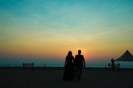 A silhouette of a couple walking hand in hand on a beach at sunset, with benches and a tent visible in the distance. The sky displays a gradient of colors from orange to blue, enhancing the peaceful and romantic ambiance.