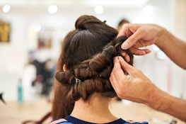 A person is styling long brown hair, creating an intricate updo with curls being pinned into place. The focus is on the hands of the stylist as they carefully arrange the hair, with a blurred salon background.
