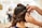 A person is styling long brown hair, creating an intricate updo with curls being pinned into place. The focus is on the hands of the stylist as they carefully arrange the hair, with a blurred salon background.